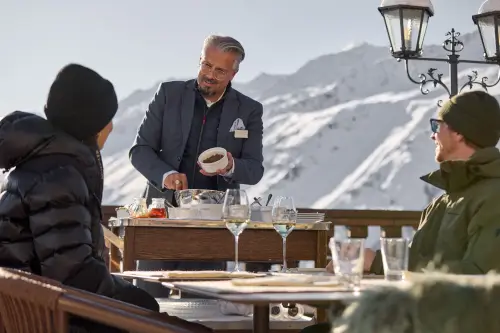 Kellner, der Gästen im Freien im TOP Hotel Hochgurgl mit schneebedeckten Bergen im Hintergrund serviert.