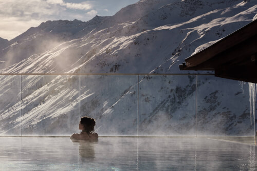 A woman swimming in an outdoor pool at TOP Hotel Hochgurgl, surrounded by majestic mountains with snow and a clear sky