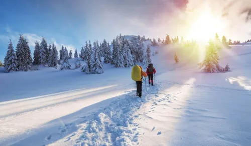 Winterwanderer in verschneiter Landschaft mit Tannenbäumen und Sonnenstrahlen.