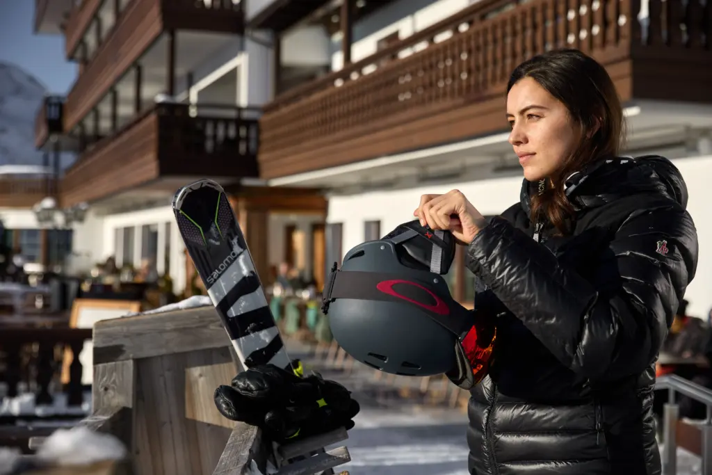 Eine Frau im Skioutfit steht mit einem Skihelm in den Händen auf der Hotelterrasse in den Bergen